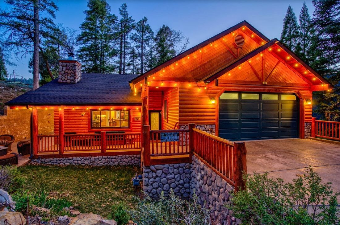 Big Bear Reset log cabin exterior at twilight with glowing string lights, stone chimney, and deck