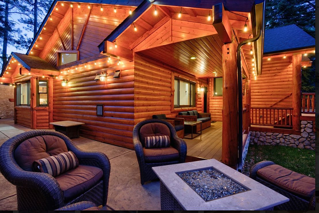 Back patio at dusk with fire pit table, wicker chairs, and string lights on the log cabin