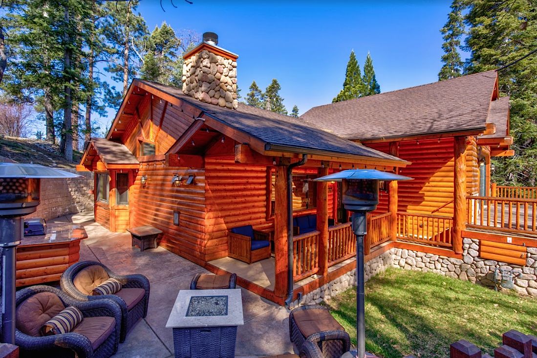 Cabin exterior with stone chimney, patio with fire pit, outdoor heaters, and pine trees